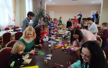 Dzieci i rodzice podczas warsztat&oacute;w, siedzą przy stołach i robią ozdoby choinkowe.