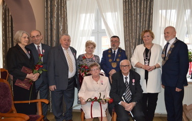 Pary pozują do zdjęcia z przedstawicielami władz samorządowych. Jedna z par siedzi na przodzie na krzesłach. W tle okna ubrane ozdobnymi firanami i zasłonami.