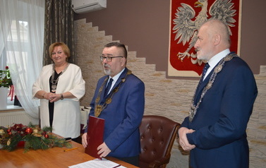 Przedstawiciele władz samorządowych (od lewej zastepca kierownika USC, burmistrz Trzemeszna i przewodniczący rady) stoją przy drewnianym stole. Burmistrz trzyma czerwoną teczkę. W tle na ścianie godło Polski.