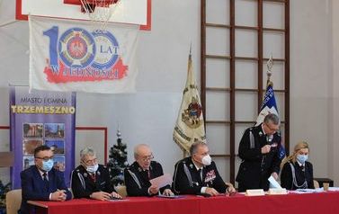 Władze OSP siedzą przy stole prezydialnym. Jeden mężczyzna stoi i przemawia. W tle sztandary strażackie i banery organizator&oacute;w.