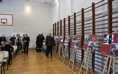 Po prawej stronie wystawa ustawiona przy barierkach gimnastycznych. Po lewej stronie uczestnicy spotkania siedzą na krzesełkach i rozmawiają.