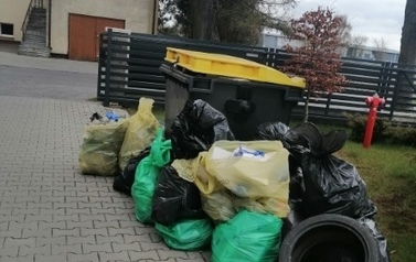 Na pierwszym tle obok pojemnika na smieci leżą worki ze śmieciami i opona. Dalej płot z poziomych paneli, a w tle fragment budynku kościoła. 
