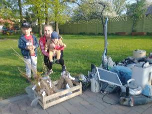 Mama zdw&oacute;jką dzieci, w tym jedno na rękach, pozują do zdjęcia. Przed nimi po lewej stronie w skrzyni sadzonki, a po prawej elektroodpady.