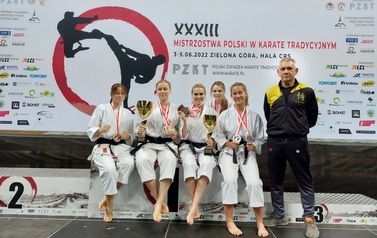 Grupa zawodniczek siedzi trzymając puchary i medale. Obok stroi trener.