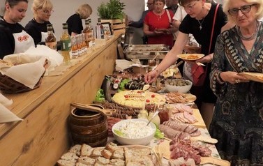 Miejsce cateringu. Po prawej zwiedzający nakładają sobie potrawy. Na środku st&oacute;ł z potrawami. Po lewej panie wydające.