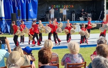 Na pierwszym planie od tyłu publiczność na krzesłach. Na drugim grupa taneczna dziewczynek podczas występu na scenie.