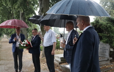 Pięciu mężczyzn stoi pod parasolami na alekce cmetranej. dw&oacute;ch trzyma zapalone znicze. Burmistrz trzyma wiązankę z biało-czerwonych kwiat&oacute;w.
