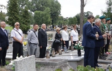 Uczestnicy wydarzenia stoją miedzy grobami. Niekt&oacute;rzy trzymają znicze.