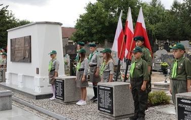 Zuchy i harcerze w mundurach stoją na warcie przy Zbiorowej Mogile Ofiar II Wojny Światowej. W tle flagi Polski i groby. 
