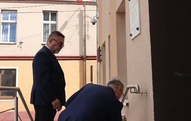 Burmistrz stoi przed tablicą burmistrza Szymańskiego, wiceburmistrz klęczy kładąc znicz.