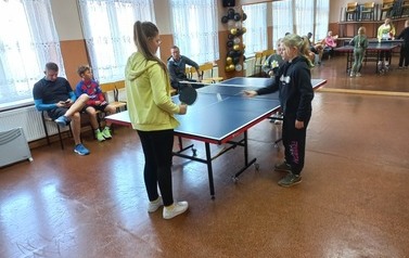 Przy stole do tenisa stoją dwie dziewczynki. W tle na krzesełkach siedzi trzech mężczyzn.