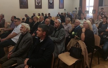 Widok na salę, gdzie na krzesełkach w kilku rzędach siedzi publiczność.