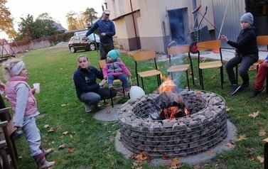 Dorośli i dzieci siedzą i stoją wok&oacute;ł ogniska.