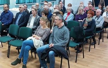 Uczestnicy spotkania siedzą na krzesełkach w auli szkoły.