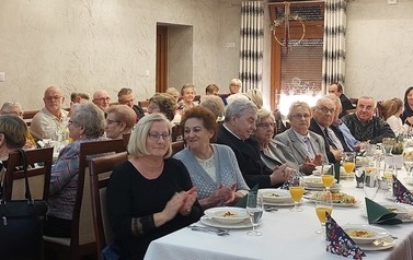 Widok na salę i siedzących przy stołach uczestnik&oacute;w spotkania. Stoły przykryte białymi obrusami, a na nich ustawiona zastawa.