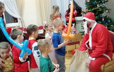 Po prawej stronie na krześle siedzi Mikołaj trzymając między stopami worek z prezentami. Po lewej kolejka dzieci z balonami. W tle choinka.