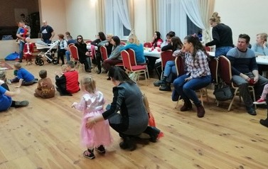 Po lewej stronie na podłodze siedzą dzieci. Po prawej rodzice siedzą przy stołach.