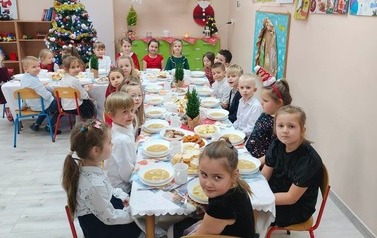 Dzieci w odświętnych strojach siedzą przy stole, na kt&oacute;rym znajdują się potrawy wigilijne.