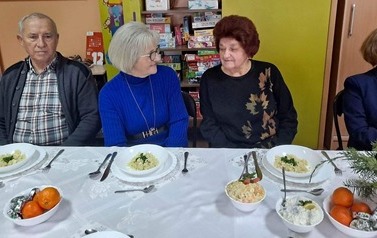 Trzy starsze kobiety i mężczyzna siedzą przy stole i rozmawiają. Na stole potrawy wigilijne.