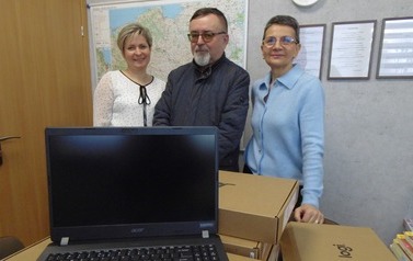 Na pierwszym planie stos karton&oacute;w z laptopami. Na g&oacute;rze jeden rozpakowany i otwarty. Za nimi stoją burmistrz, kierownik i dyrektor przedszkola nr 2.