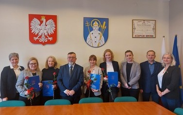 Nauczycielki, dyrektorzy, burmistrz oraz kierownik ROJO pozują do zdjęcia stojąc, w tle godło i herb gminy.