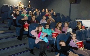 Dzieci w sali kinowej siedzą w fotelach. Niekt&oacute;re trzymają pudełka z popcornem.