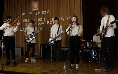 Na scenie zesp&oacute;ł muzyczny. Trzech chłopc&oacute;w gra na gitarze. Jeden na skrzypcach. Dziewczyna śpiewa przy mikrofonie na stojaku.
