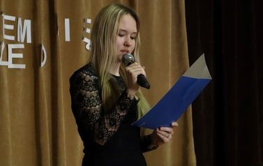 Uczennica czyta wiersz przez mikrofon. W drugiej ręce trzyma niebieska teczkę.