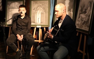 Na pierwszym planie dw&oacute;ch mężczyzn siedzących na wysokich krzesłach. Jeden śpiewa przez mikrofon. Drugi gra na gitarze.