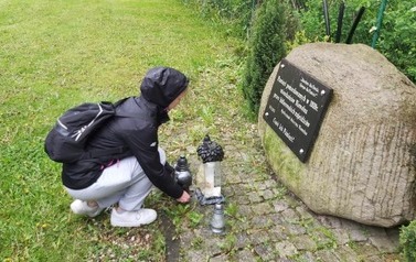 Dziewczynka kuca i kładzie znicz przy kamiennym obelisku.