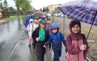 Dzieci pod parasolami i bez maszerują po chodniku. Pada deszcz.