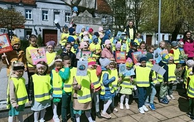 Grupa przedszkolak&oacute;w w ż&oacute;łtych kamizelkach pozuje do zdjęcia przy pomniku Jana Kilińskiego. Część trzyma w dłoniach ekologiczne transparenty.