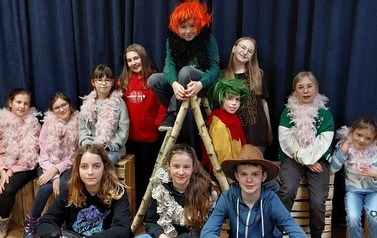 Grupa dzieci na scenie. Wszyscy siedzą na drewnianych skrzynkach, jedno na drabinie z gałęzi. Część ma na szyi r&oacute;żowe szale boa.