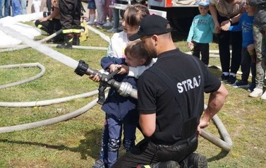 Strażak i dw&oacute;jka dzieci trzymają wąż strażacki, z kt&oacute;rego leci woda.