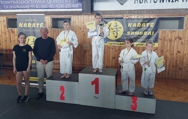 Czterech małych zawodnik&oacute;w w strojach karate stoji na podium. Na szyjach wiszą medale, a w dłoniach trzymają puchary i dyplomy. Obok podium stoją wręczający medale..