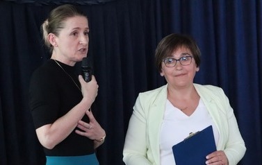 Zbliżenie na dwie kobiety. Z lewej strony z mikrofonem w ręku dyrektor szkoły. Z prawej z teczka w ręku wicedyrektor.