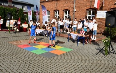Dw&oacute;ch chłopc&oacute;w w neibieskich strojach piłkarskich na kolorowej macie pokazują sztuczki z piłką. W tle reszta uczni&oacute;w biorących udział w pokazie.