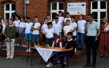 Po prawej stronie stoi dyrektor i przemawia przez mikrofon. Obok duża grupa uczni&oacute;w przygotowana do występu.