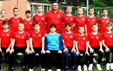 Grupa chłopc&oacute;w z trenerem w czerwono-czarnych strojach pozuje do zdjęcia.