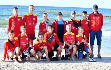 Grupa chłopc&oacute;w z trenerem w czerwonych bluzach pozuje do zdjęcia na plaży. W tle morze.
