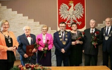 Dwie pary oraz wręczający medale pozują do zdjęcia na tle godła Polski