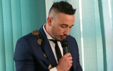 Mężczyzna z insygniami na szyi stojąc przemawia przez mikrofon.