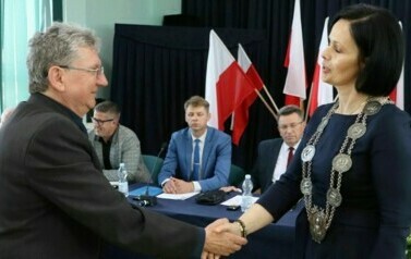 Kobieta z insygniami na szyi gratuluje mężczyźnie. W tle siedzą radni.