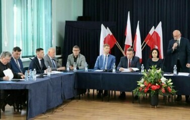 Siedmiu mężczyzn i kobieta siedzi przy stole ustawionym w literę L, jeden mężczyzna stoi i przemawia przez mikrofon. Za nimi statyw z kilkoma flagami Polski.