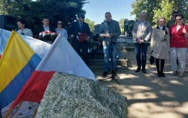 Na pierwszym planie z lewej strony gragment obelisku oraz powiewające flagi gminy i Polski. Na drugim rząd dorosłych os&oacute;b. Mężczyzna przemawia przez mikrofon. W tle nagrobki.