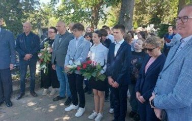 Grupa os&oacute;b w r&oacute;żnym wieku. Niekt&oacute;re osoby trzymają biało-czerwone wiązanki.