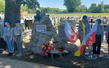 Na pierwszym planie obelisk z kamieni, przed nim stojak z flagami Polski i Gminy Trzemeszno. Po bokach stoją po trzy dziewczynki &ndash; zuchy. W tle nagrobki.