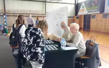 Przy stoliku siedzi kobieta i mężczyzna. Z drugiej strony stoi grupa dzieci.