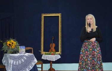 Autorka przemawia na scenie. Z lewej strony rama od obrazu ze skrzypcami w środku oraz wazon z kwiatami/
