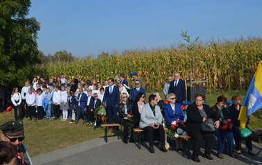 Na pierwszym planie uczestnicy spotkania siedzą na ławkach, w tle stoją uczniowie
